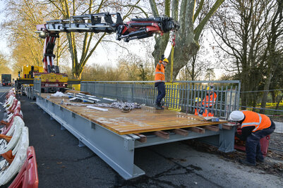 Ouvriers en tenue de chantier guidant la pose par grue de la passerelle métallique à platelage bois