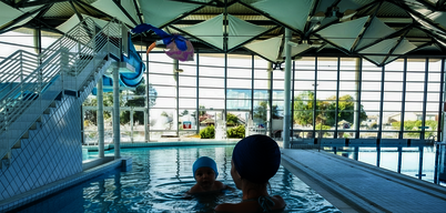 Un adulte accompagne un nourrisson dans le bassin intérieur d'un centre aquatique couvert, avec jouets gonflables colorés au-dessus de l'eau