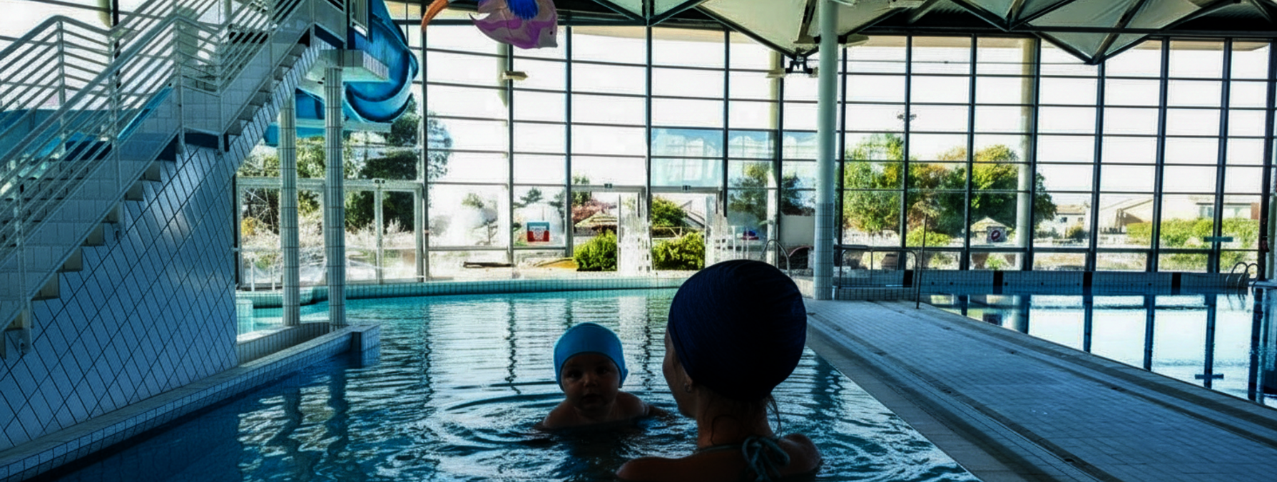 Un adulte accompagne un nourrisson dans le bassin intérieur d'un centre aquatique couvert, avec jouets gonflables colorés au-dessus de l'eau