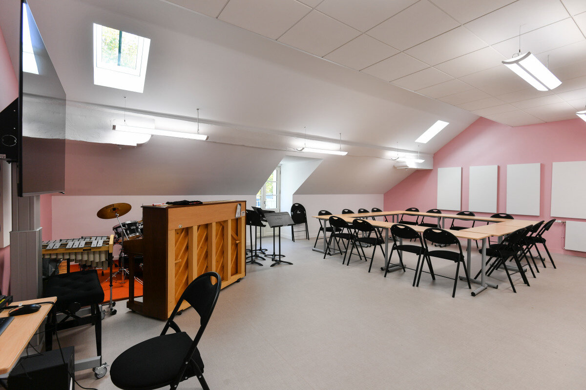 Salle de cours équipée d'un piano, d'une batterie et d'un marimba, avec tables disposées en U