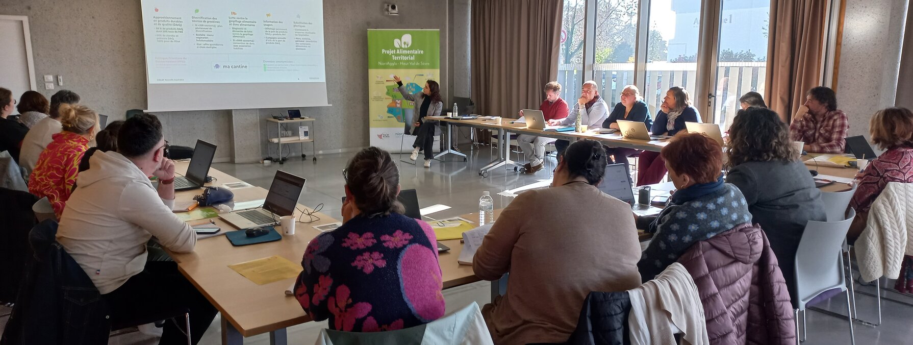 Salle de réunion avec une vingtaine de participants munis d'ordinateurs face à un écran de projection et une bannière Projet Alimentaire Territorial.