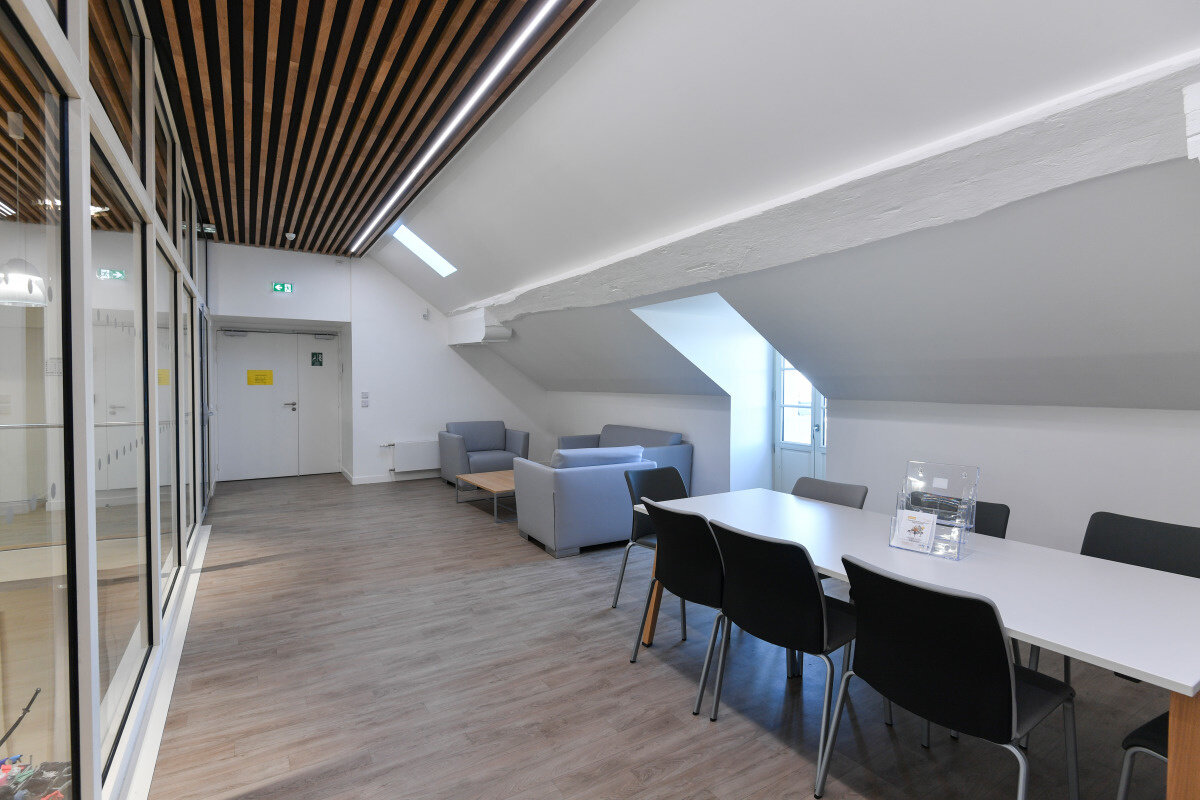 Salle commune avec table et chaises noires, canapés gris et cloison vitrée sous poutre de charpente apparente