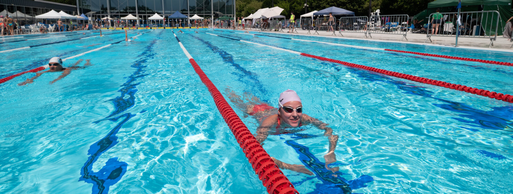 Nageurs dans les couloirs du bassin extérieur en plein soleil, avec le bâtiment de la piscine en fond