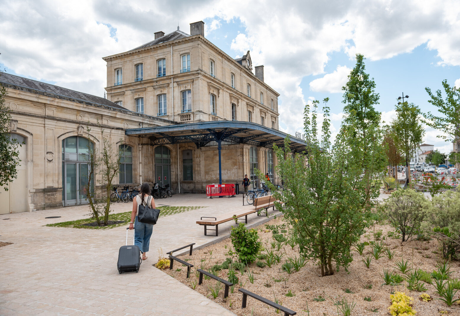 Parvis réaménagé avec arbres, végétation et bancs en bois, un voyageur tirant une valise vers l'entrée de la gare - Agrandir l'image 1 sur [1], fenêtre modale