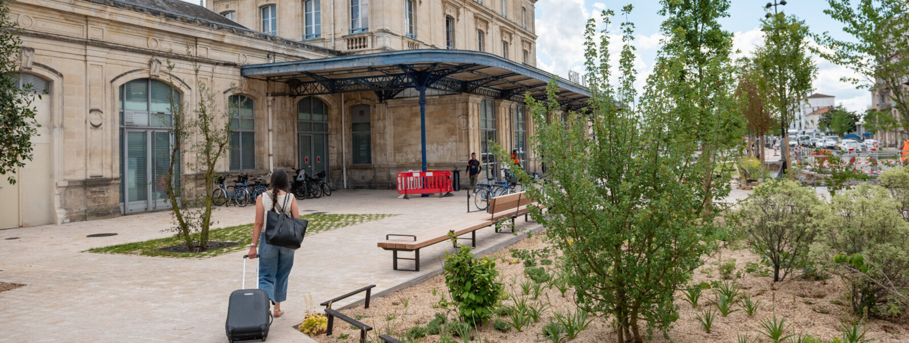 Parvis réaménagé avec arbres, végétation et bancs en bois, un voyageur tirant une valise vers l'entrée de la gare