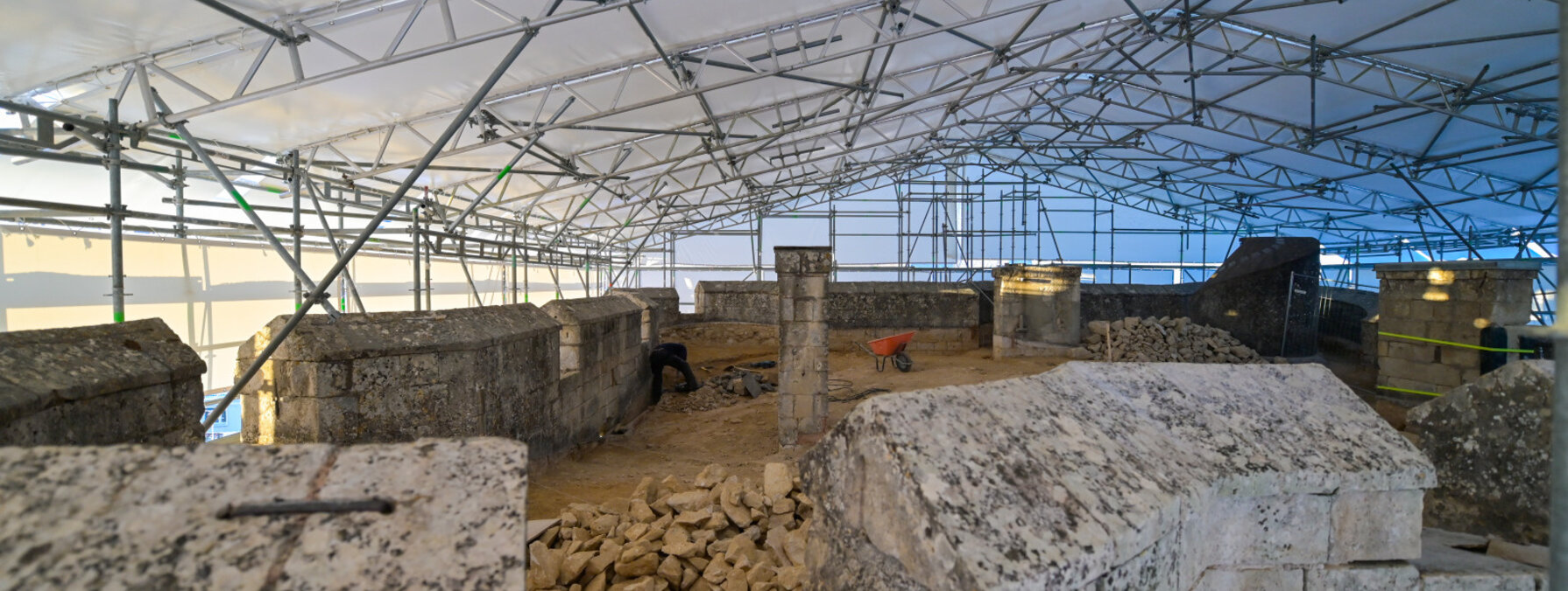Plateforme sommitale couverte d'une tente de chantier sur échafaudages métalliques, blocs de tuffeau au sol et artisan à l'œuvre