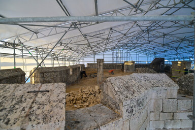 Plateforme sommitale couverte d'une tente de chantier sur échafaudages métalliques, blocs de tuffeau au sol et artisan à l'œuvre