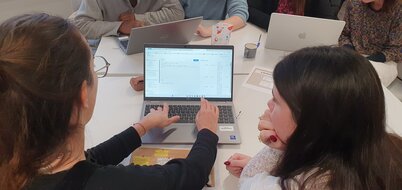 Femmes réunies autour d'ordinateurs portables lors d'un atelier collaboratif, l'une désignant une interface à sa voisine