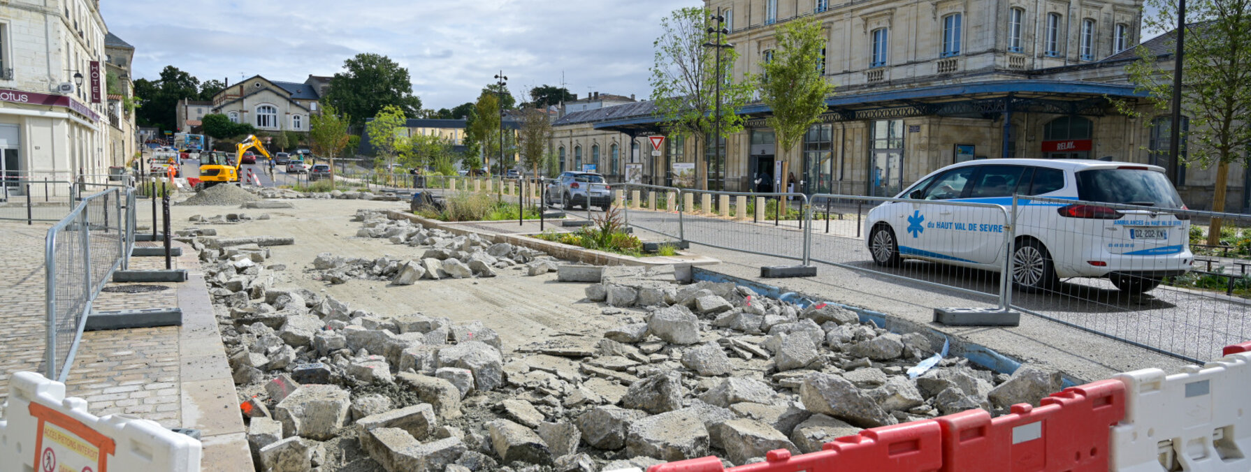 Dalles de béton brisées au sol et barrières de sécurité rouge et blanc délimitant le chantier, façade de la gare en arrière-plan.