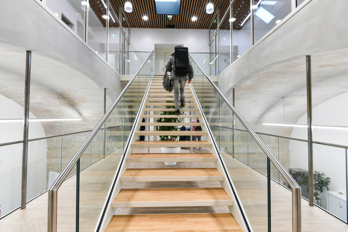 Hall du Conservatoire, escalier central à rampes vitrées, un élève portant un étui d'instrument monte les marches