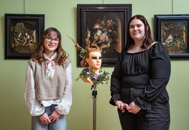 Deux apprenties coiffeuses posent dans une salle du musée encadrant une tête à coiffer ornée d'un chignon fleuri élaboré