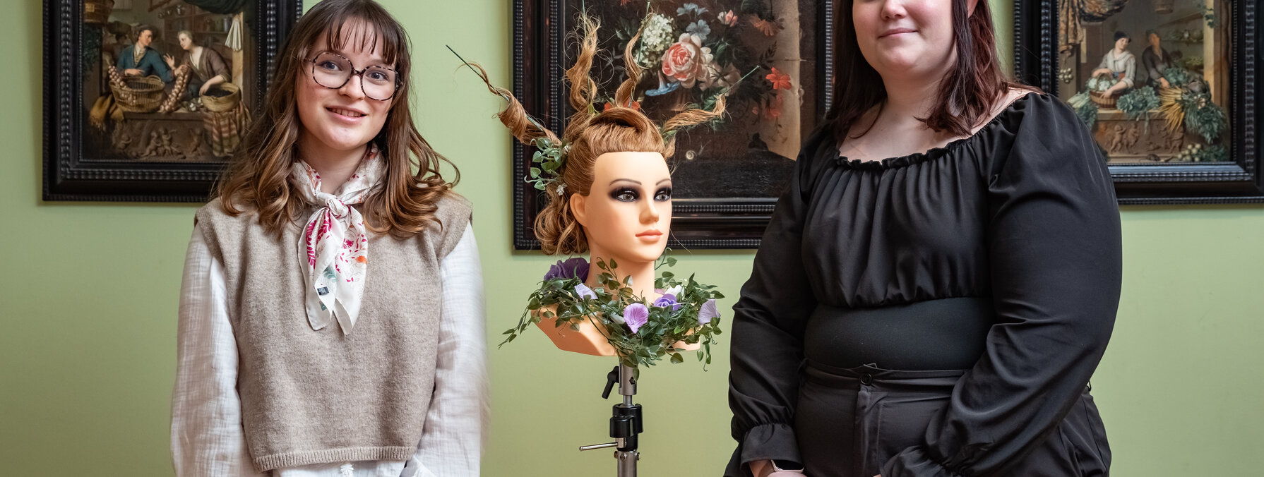 Deux apprenties coiffeuses posent dans une salle du musée encadrant une tête à coiffer ornée d'un chignon fleuri élaboré