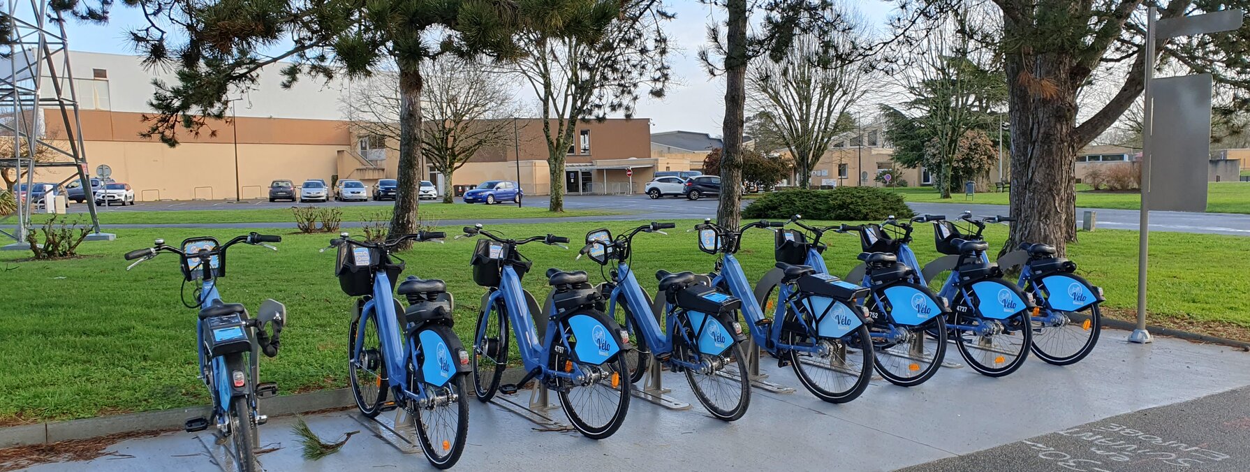 Sept vélos bleus en libre-service alignés en rang sur un emplacement balisé, avec une borne de déverrouillage et des espaces verts en arrière-plan