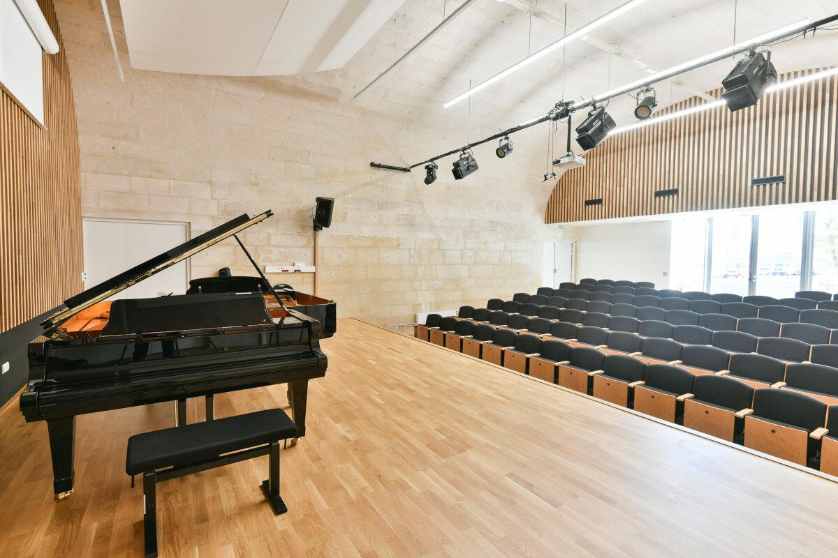 Salle de concert du conservatoire avec piano à queue sur scène en parquet, gradins vides et projecteurs de scène
