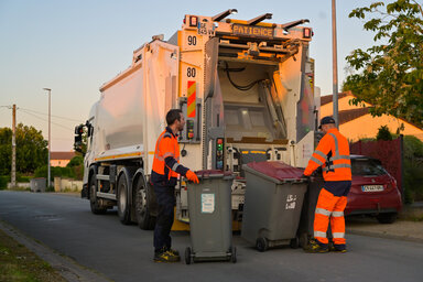 Deux agents de collecte manœuvrent des bacs gris devant un camion-benne dans une rue résidentielle