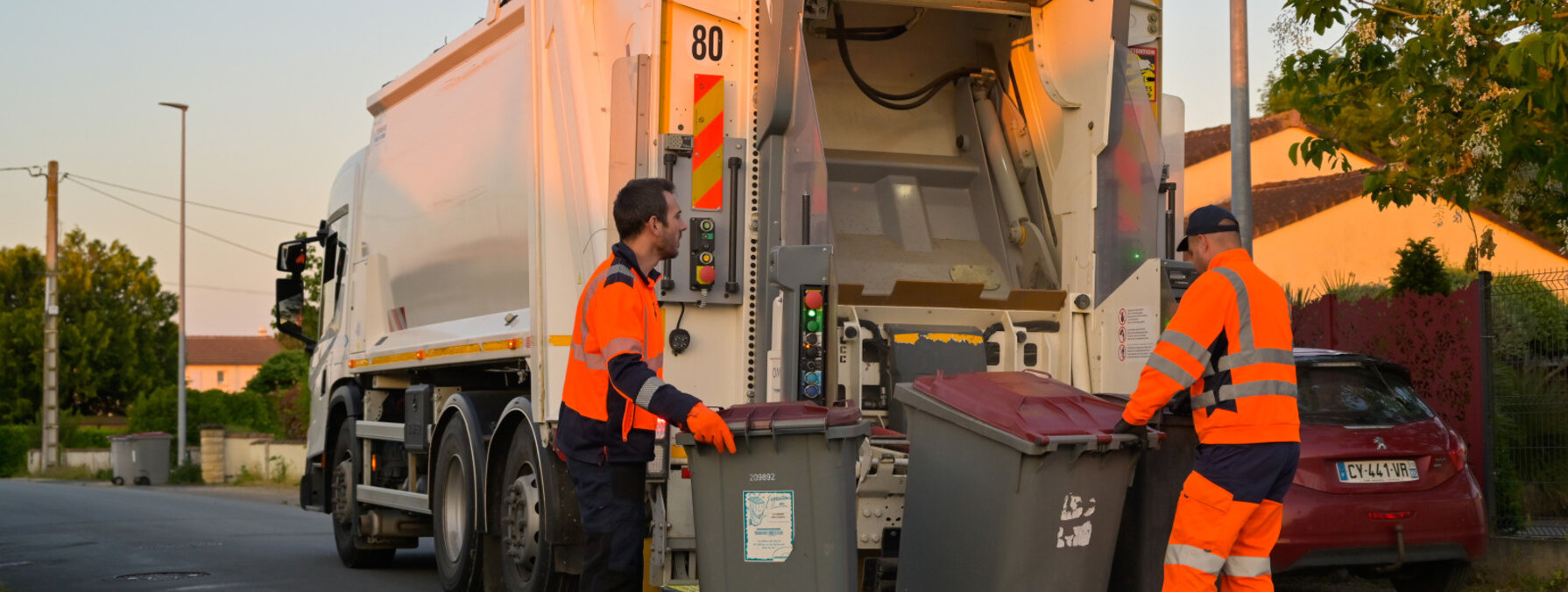 Deux agents de collecte manœuvrent des bacs gris devant un camion-benne dans une rue résidentielle