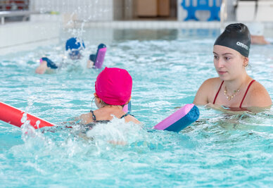 Monitrice en bonnet Arena accompagnant un enfant avec frite et planche dans un bassin couvert